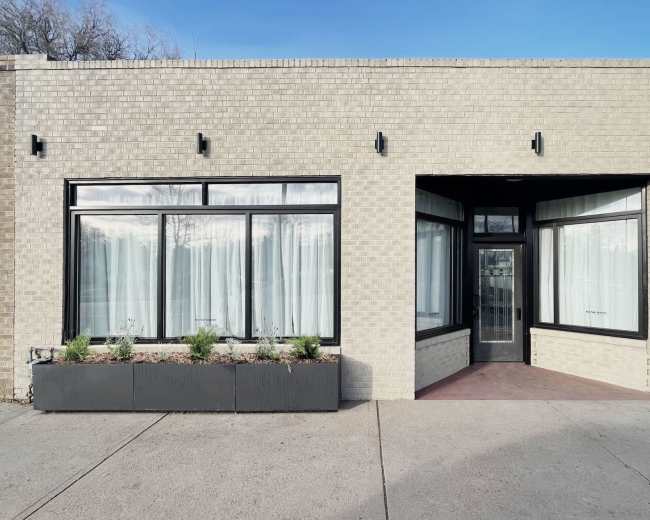 The image shows a single-story brick building with large front windows and a centrally located door, flanked by a planter box containing greenery.