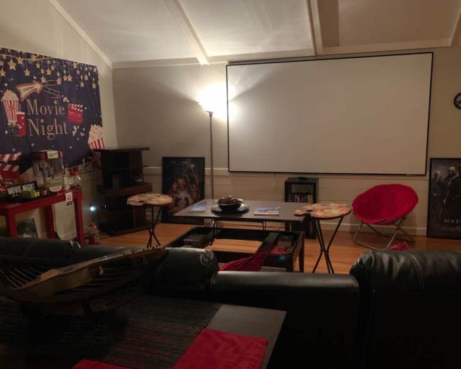 The image shows a cozy home theater setup with a large projector screen, seating area, and movie-themed decorations including popcorn and film posters.
