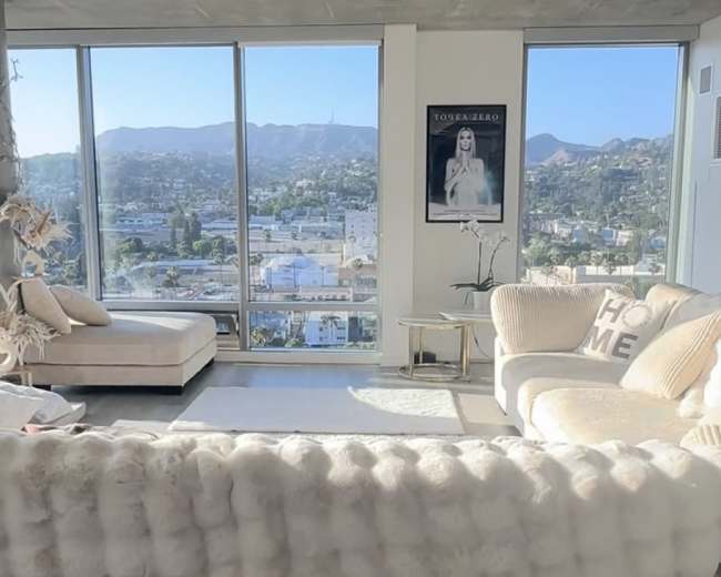 A bright living room features a large sectional couch, a chaise lounge, and large windows showcasing a mountainous cityscape.