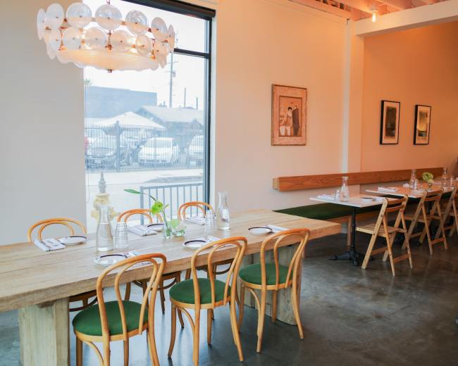 The image shows a modern restaurant interior featuring a long wooden table with green chairs, a large window, and simple decor.