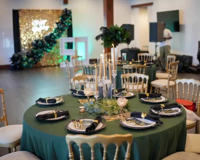 The image shows a beautifully arranged dining table with green tablecloth, elegant gold chairs, and decorative items, set in a well-lit event space adorned with a backdrop of balloons and plants.