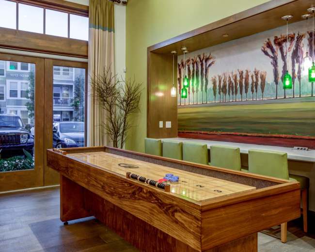 A wooden shuffleboard table is placed in a brightly lit room with large windows, featuring a painted landscape mural and a row of green barstools.