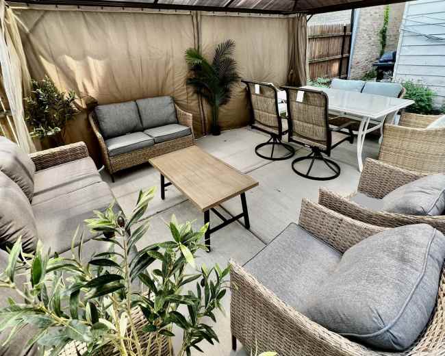A furnished outdoor gazebo features seating arrangements, a central table, and potted plants.