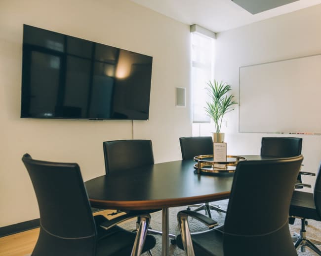 A modern conference room features a large television, a whiteboard, a round table, and six black chairs arranged around it, with a small decorative plant in the center.