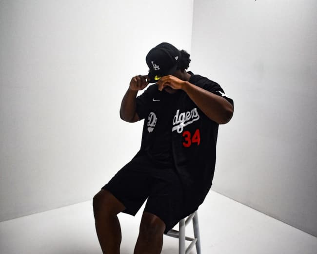 A person wearing a Dodgers jersey and cap sits on a stool in a plain, well-lit studio.