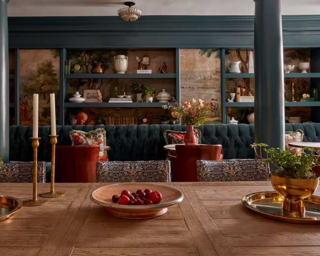 A dining area features a wooden table with decorative bowls and candles, surrounded by upholstered chairs and shelves displaying various decor items.