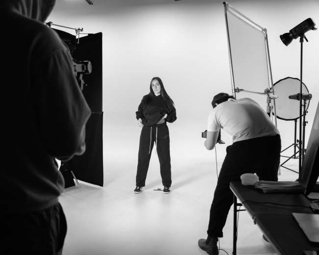 A person in a hooded outfit poses for a photo while another individual sets up a camera in a studio.