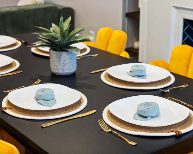 A dining table is set with six white plates on woven placemats, adorned with rolled blue napkins and a succulent centerpiece, surrounded by yellow chairs.