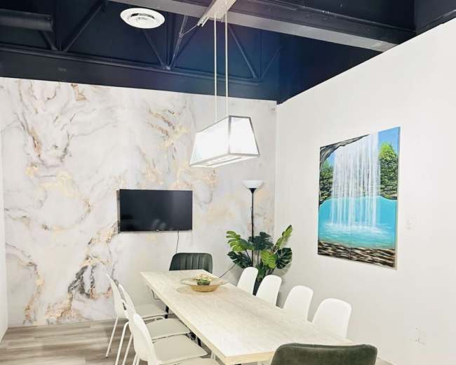 A conference room with a long table surrounded by white and green chairs, featuring a large painting of a waterfall on the wall and a television mounted nearby.