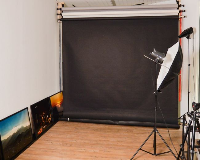 The image shows a photography studio with a black backdrop setup, a light source on a stand, and framed photos leaning against the wall.