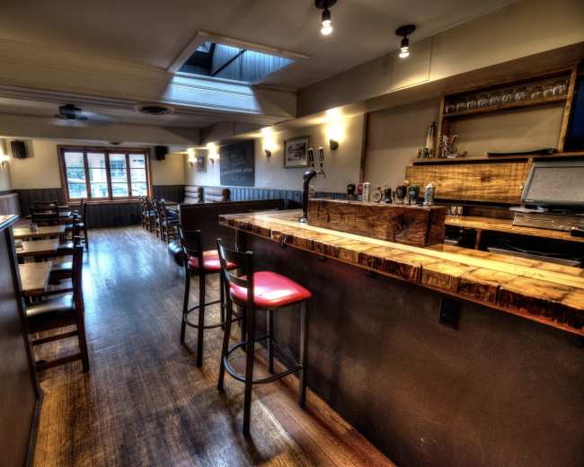 The image depicts the interior of a bar featuring wooden furniture, a polished bar counter, and multiple seating areas illuminated by soft lighting.