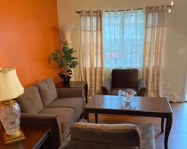 A living room furnished with a brown sofa, a pair of armchairs, a coffee table, and a lamp, alongside a window with sheer curtains.