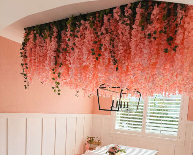 A dining area features a long table with pink chairs beneath a floral installation of pink flowers hanging from the ceiling.