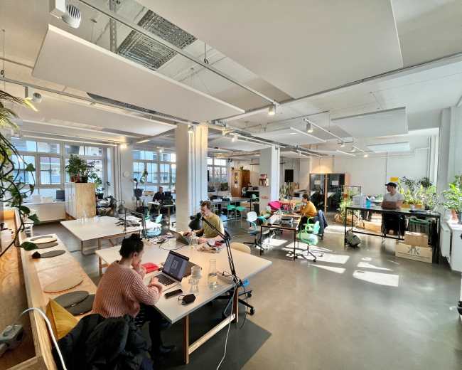 The image shows a spacious office environment with multiple workstations, natural light streaming in through large windows, and several people engaged in various tasks.