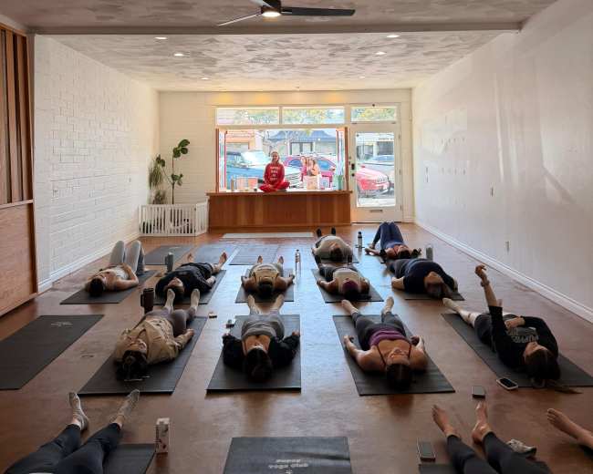 The image shows a group of people lying on yoga mats in a studio with a person standing at the front near a counter.