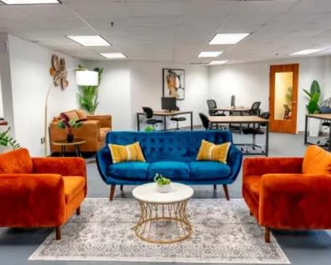 The image shows a modern office lounge area featuring a blue sofa flanked by orange armchairs, a round coffee table, and workspace desks in the background.