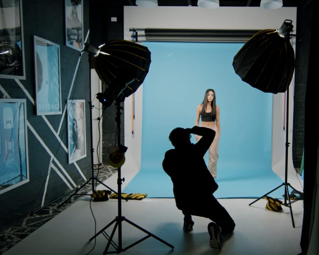 A photographer is capturing images of a model against a blue backdrop in a studio setup with lighting equipment.