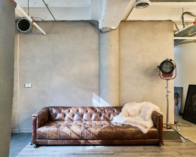 The image features a brown leather sofa against a plain, light gray wall with a vintage studio light positioned nearby.