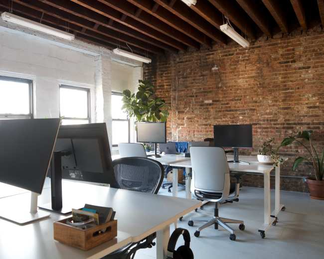 The image shows a modern office space with several desks and monitors, plants, and exposed brick walls.
