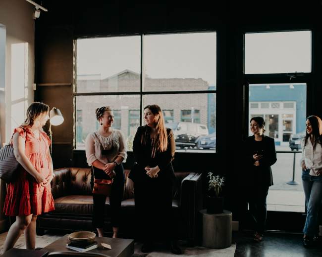 A group of women stands conversing in a modern, well-lit coffee shop with large windows.