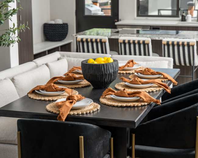 A dining table is set with plates and brown napkins, surrounded by black chairs, and a bowl of lemons sits in the center.