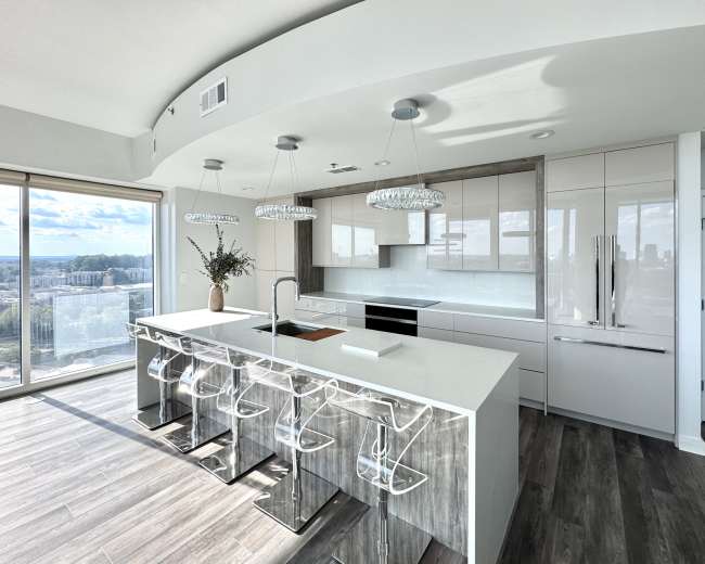 The image shows a modern kitchen with a large island, sleek cabinetry, and several bar stools, overlooking a city skyline through large windows.