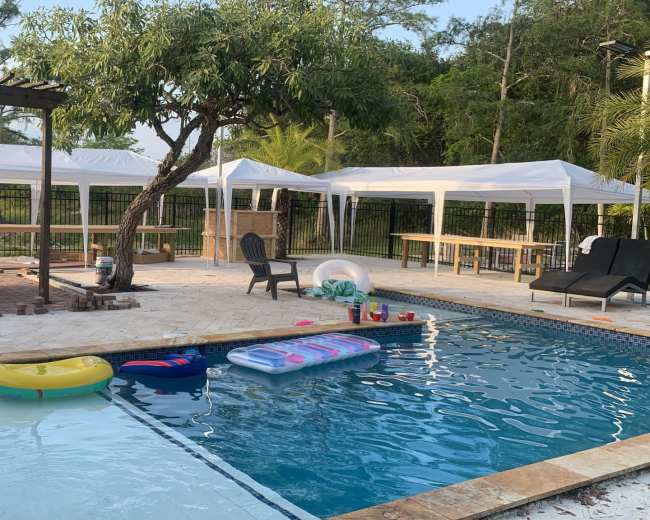 The image shows a backyard pool area with floating pool toys, lounge chairs, and white canopies set up nearby.