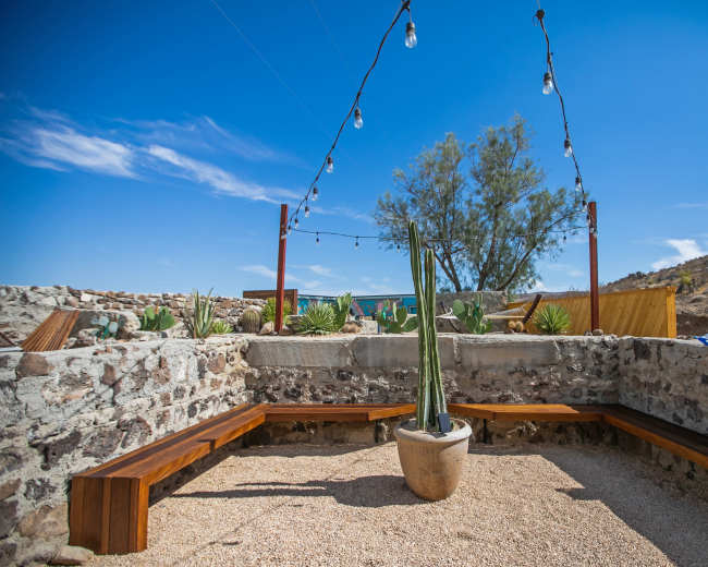 A circular seating area made of stone and wood surrounds a potted plant under string lights, set against a desert landscape.