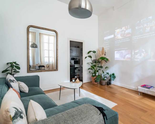 A bright living room features a green couch, a large mirror, and a projection on the wall displaying various media content.