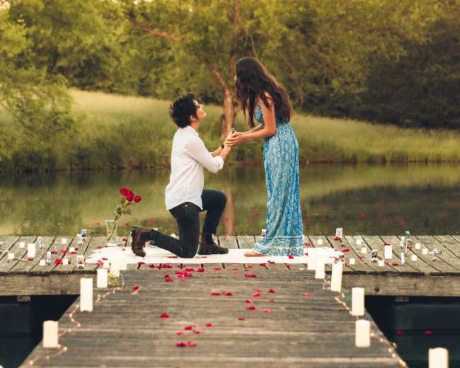 A man proposes to a woman on a dock, surrounded by rose petals and candles.