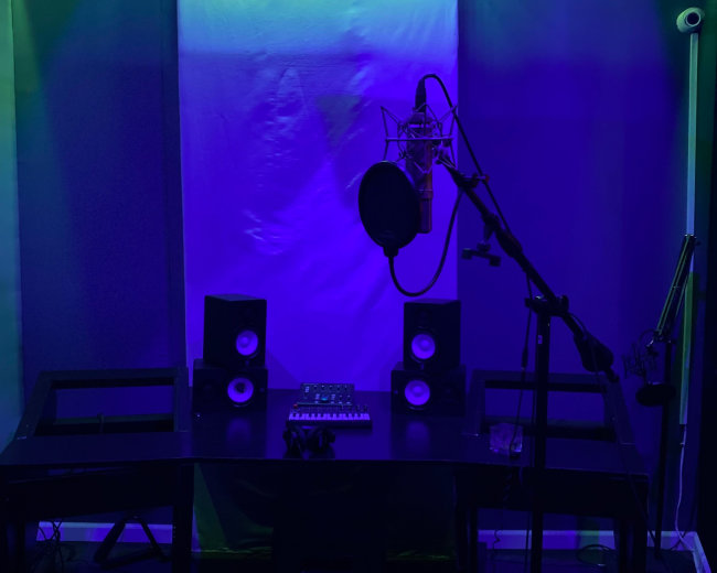 A dimly lit recording studio features a microphone, speakers, and a sound mixer on a black desk, surrounded by green and blue lights.