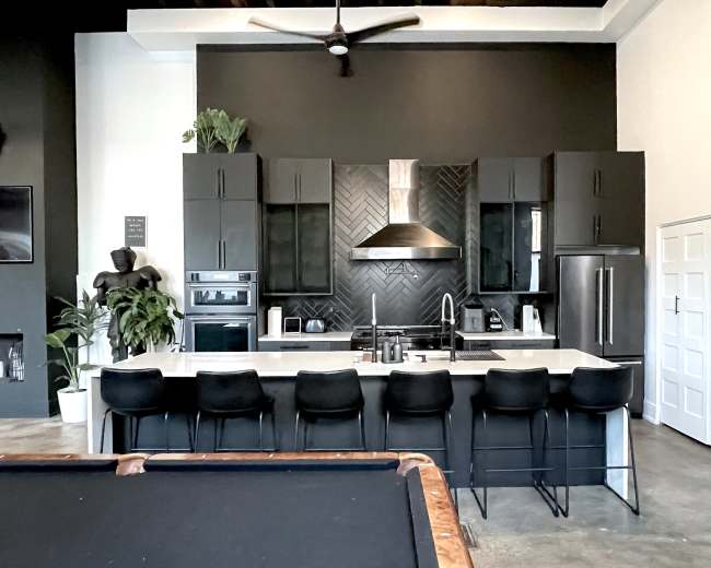 The image shows a modern kitchen with black cabinetry, a white countertop, and a large island with several black bar stools.