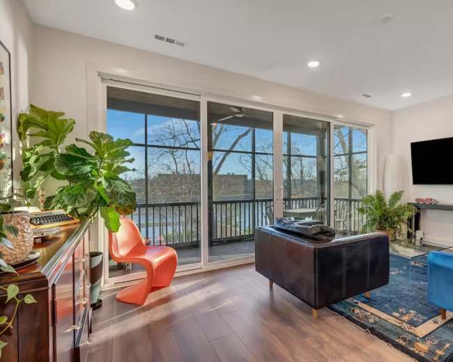 The image shows a modern living room with large windows overlooking a river, furnished with a blue couch, a black lounge chair, and a bright orange chair.