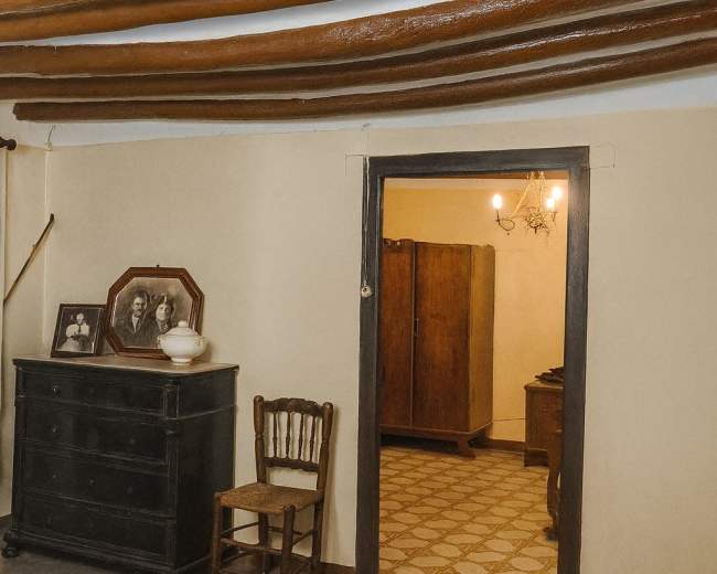 The interior of a simple room features a wooden chair and a dark dresser with framed photographs, leading to a second room with a wardrobe and a light fixture.