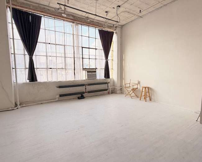 The image shows a spacious, empty studio with large windows, wooden flooring, and a few simple pieces of furniture.