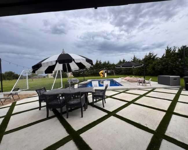 The image shows a patio area with a dining table, black and white umbrellas, a swimming pool, and a grassy lawn in the background.
