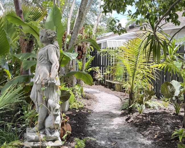 A stone statue stands next to a winding path surrounded by lush greenery and tropical plants.