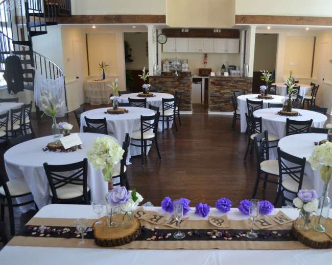 A decorated event space with circular tables arranged around a central bar area, featuring floral centerpieces and a wooden decorative runner.