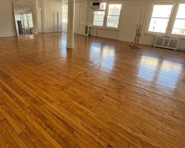 The image shows a spacious, empty room with polished wooden floors, large windows, and white walls.