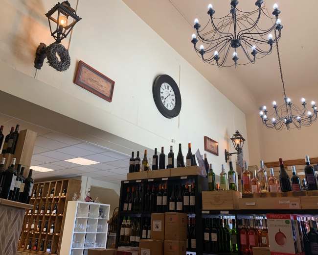 A wine shop interior with shelves of bottles, a clock on the wall, decorative lamps, and a chandelier overhead.