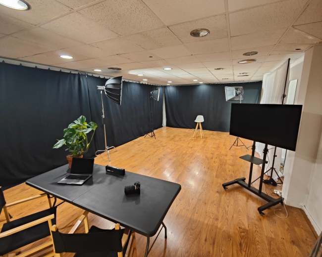 The image shows a spacious photography studio featuring black backdrop curtains, a wooden floor, a table with equipment, and various lighting setups.