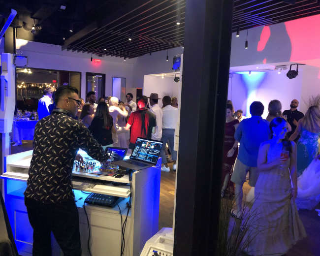 A DJ mixes music at a lively dance party in a modern venue filled with guests dancing and socializing.