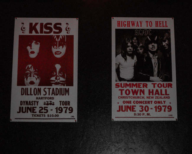 A black and white checkered skateboard mounted on a wall above two vintage concert posters for the bands Kiss and AC/DC, along with a red star light.
