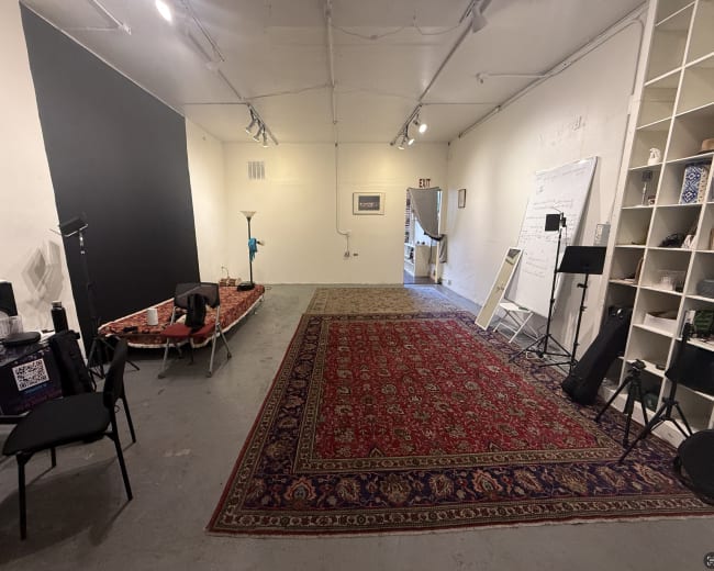 The image shows a spacious, well-lit room with a large area rug, a black wall section, and various furniture items including chairs and a lamp.