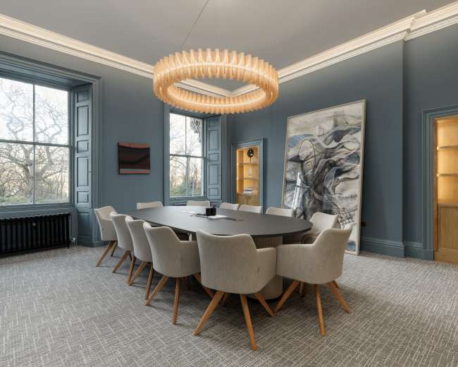 A modern conference room features a large oval table surrounded by upholstered chairs, illuminated by a distinctive circular chandelier, with large windows providing natural light.