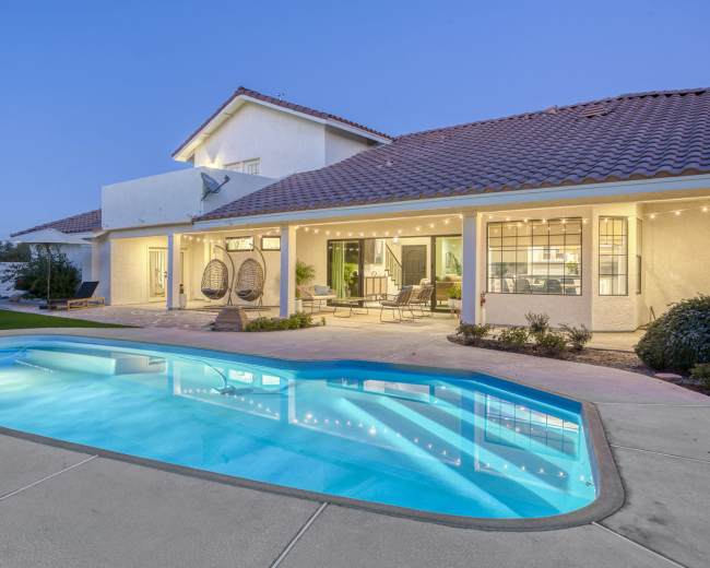 A two-story house features a pool in the backyard with hanging chairs and patio seating under string lights.