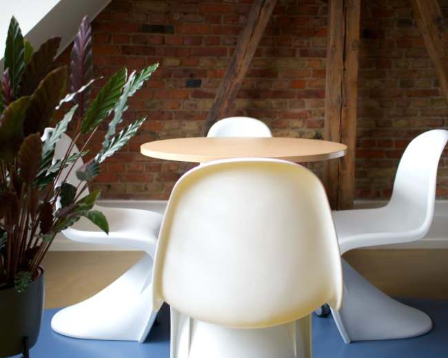 A round wooden table is set in a room with exposed brick walls, accompanied by three white chairs and a potted plant in the foreground.