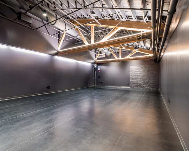 The image shows an empty industrial-style room with a polished concrete floor, dark walls, and exposed wooden beams in the ceiling.