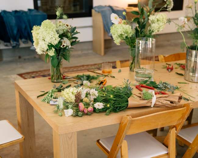 A wooden table is surrounded by folding chairs, with various flowers and greenery scattered across the surface alongside gardening tools and a bag.