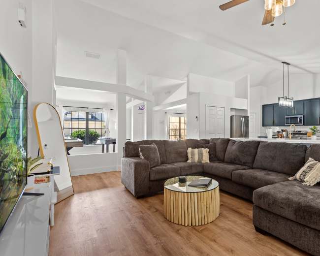 A spacious living room features a large sectional sofa, a round wooden coffee table, and a view of an open kitchen with dark cabinetry.